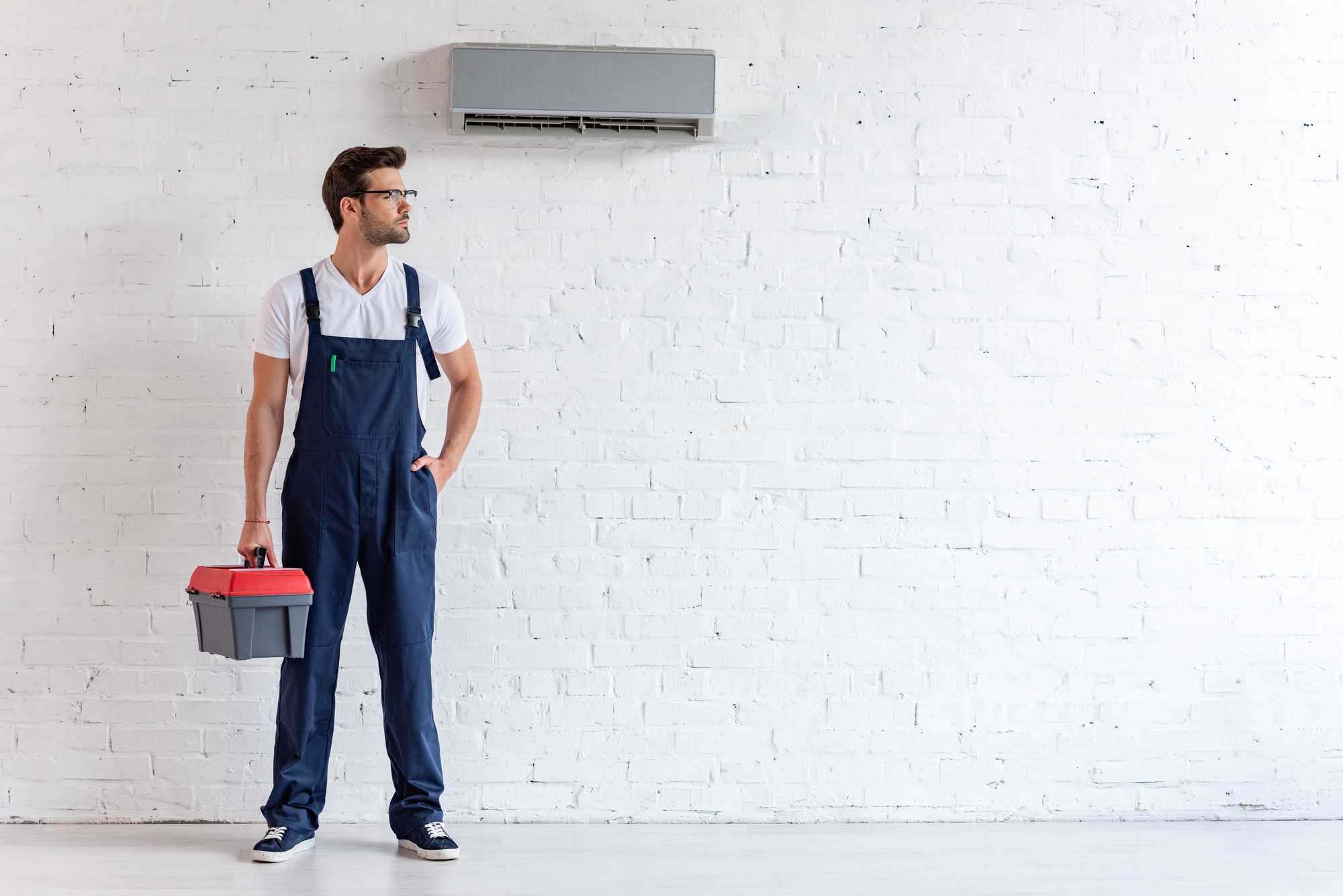 handsome repairman in uniform standing under conditioner and looking away